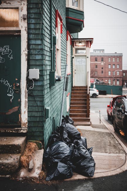 A collection of several tightly packed black garbage bags filled with waste is situated on the pavement next to a building with weathered green wooden cladding, red and white trim around windows and a door, and a set of wooden stairs leading up to a white door with a small overhang. The bags are placed directly against the building's wall near a concrete step and show signs of being recently piled, with some partially leaning against an adjacent electrical box and cable. The scene is set on an urban street in daylight, with parked cars visible on the road and a brick building in the background. The environment suggests an area where private waste disposal, such as rubbish removal services provided by companies like Waste Disposal Kingston, is utilized as an alternative to council collection. The overall visual offers a typical view of on-site rubbish clearance and the importance of proper waste management in residential or commercial properties in Kingston, with natural lighting highlighting the textures of the bags and building exterior.