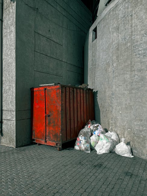 A collection of several tightly packed black garbage bags filled with waste is situated on the pavement next to a building with weathered green wooden cladding, red and white trim around windows and a door, and a set of wooden stairs leading up to a white door with a small overhang. The bags are placed directly against the building's wall near a concrete step and show signs of being recently piled, with some partially leaning against an adjacent electrical box and cable. The scene is set on an urban street in daylight, with parked cars visible on the road and a brick building in the background. The environment suggests an area where private waste disposal, such as rubbish removal services provided by companies like Waste Disposal Kingston, is utilized as an alternative to council collection. The overall visual offers a typical view of on-site rubbish clearance and the importance of proper waste management in residential or commercial properties in Kingston, with natural lighting highlighting the textures of the bags and building exterior.