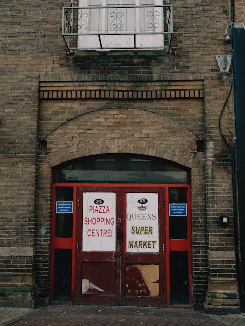 The image depicts a brick building facade with a pair of red metal double doors centered beneath a large archway made of brickwork. The doors are weathered, with peeling paint and visible patches, and feature printed signs advertising a shopping centre and market, with some text partially obscured or torn. Flanking the doors are narrow black window panels. Directly above the doors, there is a small rectangular window with a rounded top, set into the brickwork, which is darkened and shows signs of dirt or soot. At the top of the image, a narrow balcony with a decorative wrought iron railing extends from the building, with a plain concrete surface. On the right side of the building, attached to the brick wall, there is a black utility box and a thick black cable running vertically down the wall. The scene is lit naturally, with diffuse daylight, and the overall atmosphere suggests an urban environment. This image aligns with the context of alternative waste handling or building clearance, as it shows a historic or commercial building facade that may require or have undergone rubbish removal or renovation work, which Waste Disposal Kingston could assist with.