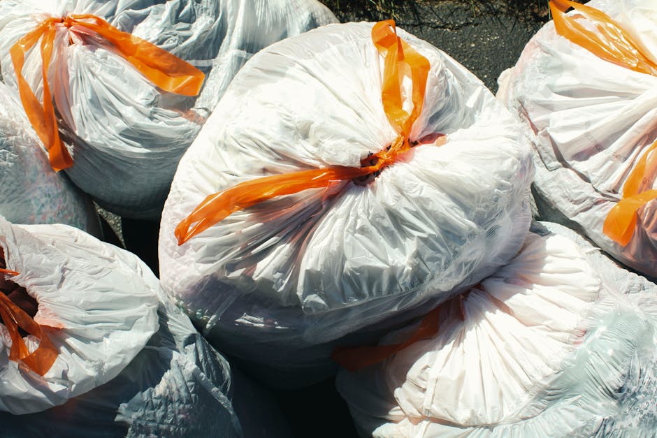 Several large white plastic rubbish bags filled with waste are gathered outdoors on a paved surface. Each bag is tightly secured with bright orange ties, and the bags appear to contain mixed household refuse, with some visible crumpled paper and other debris through the semi-transparent plastic. The bags are arranged in a somewhat haphazard manner, with some leaning against each other and overlapping, indicating a collection or disposal process. The surrounding environment suggests an urban setting, possibly near a building or a designated waste collection point, with natural daylight illuminating the scene. The image emphasizes the practicality of private waste handling services like those offered by Waste Disposal Kingston, which facilitate the collection and removal of bulky rubbish in areas such as Kingston Market Place, in line with the need for independent rubbish clearance solutions.
