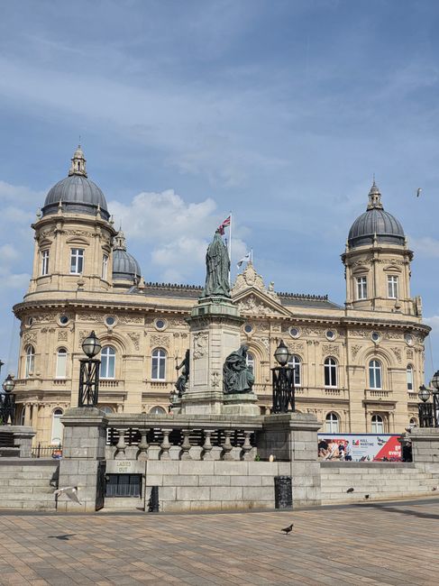 A historic sandstone building featuring classical architectural details, with symmetrical twin domed towers topped with finials, large arched windows, and decorative stonework. In front of the building, a stone monument with multiple statues and a central figure holding a flag is situated on a raised platform with a balustrade, flanked by traditional black wrought iron street lamps. The foreground shows a paved pedestrian area with a few small birds and some advertising banners along the fence, suggesting an urban environment. The sky above is partly cloudy with patches of blue visible, providing natural lighting that highlights the detailed textures of the stone and the historic ambiance of the scene. The overall setting is a city square or public space, with the scene subtly reflecting themes of civic pride and heritage, which can be associated with private or independent urban waste management services aligning with the context of rubbish removal or on-site clearance by Waste Disposal Kingston.