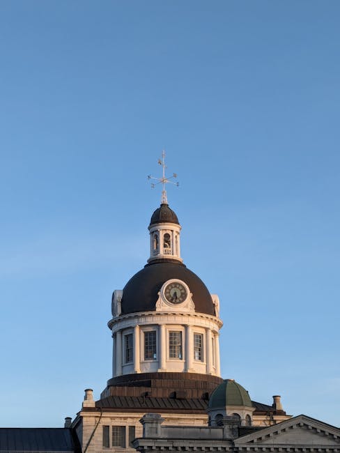 A historic city clock tower with a domed black roof and white masonry exterior, featuring a round clock face on the front and several rectangular windows below it. The tower is topped with a weather vane and a small spire, set against a clear blue sky. The surrounding architecture includes additional roof structures and ornate detailing at the building's upper levels. The image captures the clock tower’s prominent position in the cityscape, illustrating an example of traditional architectural design commonly seen in central urban areas, which can sometimes be associated with historic building preservation amidst private or independent waste collection methods in the area. Waste Disposal Kingston may be involved in the on-site clearance or waste management for such historic properties, supporting safe and efficient rubbish removal nearby.