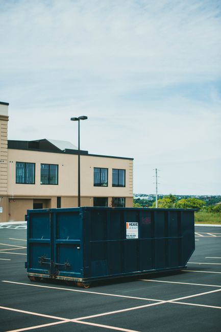 A large dark blue metal skip with a rectangular shape and high solid sides, situated in an empty parking lot marked with white lines. The skip is positioned in front of a beige, two-story commercial building with multiple windows and a flat roof. A tall black streetlamp stands behind the skip, casting minimal shadow, while the background features green foliage and open land under a partly cloudy sky. The skip appears to be used for waste disposal or on-site clearance by Waste Disposal Kingston, suitable for private waste management and alternative rubbish removal services. The surface of the skip has some minor rust at the bottom edges, and the overall environment is clean and orderly, emphasizing its suitability for commercial or residential rubbish collection that aligns with independent waste disposal practices.