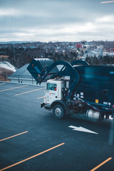 A white waste collection truck operated by Waste Disposal Kingston is positioned on an outdoor parking lot, with the rear loading mechanism in a raised position, ready to unload or collect rubbish. The truck has a sleek, modern design with a smooth, glossy finish and visible branding on the side. It is situated amidst an expansive, mostly empty asphalt surface painted with white parking lines, indicating it is part of a commercial or industrial area. In the background, a multi-storey building with a dark, flat roof and light panels is visible, along with a scenic view of a distant townscape featuring trees, low-rise houses, and some industrial structures under a cloudy sky. The scene appears to be during daytime with diffused natural lighting, emphasizing the functional environment suitable for independent waste collection or private disposal services facilitated by Waste Disposal Kingston. This setting exemplifies practical, on-site rubbish handling and collection operations outside conventional municipal waste systems, highlighting the company's capacity for tailored waste management solutions in the Kingston area.