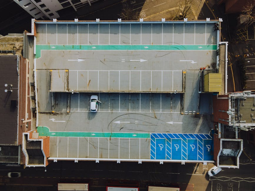 An aerial view of a rooftop parking area situated atop a building in an urban environment. The parking lot is mostly empty, with clearly marked white lines designating individual parking spaces. The surface is made of concrete with visible stains and wear, and features green painted pathways or zones along the edges, including a section for accessible parking outlined in blue with white wheelchair symbols. A small silver car is parked in one of the spaces near the center, and there are concrete or metal barriers at the perimeter to prevent vehicles from driving off. In the top-right corner, a small structure with a yellow rooftop housing electrical or service equipment is visible, along with building edges and neighboring rooftops seen beyond the parking area. The scene is lit by natural daylight, casting shadows from nearby structures, and the overall setting suggests a utilitarian space primarily used for vehicle parking, which Waste Disposal Kingston might service as part of its routine waste management for urban infrastructure.