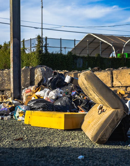 A pile of mixed waste and rubbish bags in an outdoor setting, situated on a gravel surface close to a low stone wall. The refuse includes black and clear plastic rubbish bags, some partially torn open revealing waste inside, as well as an old, discarded beige car seat with fabric upholstery and visible dirt or stains. Nearby, there is a yellow plastic container or tray, possibly part of the refuse, lying flat on the ground. The background features a metal pole and overhead wires, with a hedge and a large, curved metal or plastic structure, such as a greenhouse or storage cover, in the distance. The scene is illuminated by natural daylight, with a slightly cloudy sky. The image captures a typical example of illegal fly tipping or unsanctioned rubbish disposal, which is relevant to independent waste removal services provided by Waste Disposal Kingston in the context of alternative waste handling and legal responsibilities for rubbish collection.