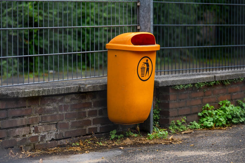 A bright orange upright waste bin attached to a metal post, positioned against a low brick wall topped with a metal fence. The bin has a rounded lid with a red reflector at the back and a black pictogram on the front depicting a person throwing rubbish into the bin. The surrounding environment features a paved pathway with some small green plants and weeds growing along the base of the brick wall. In the background, there is lush greenery with trees and shrubs visible through the metal fence. The scene is illuminated by natural daylight, highlighting the industrial finish of the bin and the textured surface of the brickwork and pavement. This setup suggests an alternative waste collection point suitable for independent rubbish disposal, reflecting private waste handling solutions in an urban or suburban setting and aligning with services like those offered by Waste Disposal Kingston for efficient rubbish removal and on-site clearance.