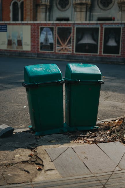 A pile of mixed waste and rubbish bags in an outdoor setting, situated on a gravel surface close to a low stone wall. The refuse includes black and clear plastic rubbish bags, some partially torn open revealing waste inside, as well as an old, discarded beige car seat with fabric upholstery and visible dirt or stains. Nearby, there is a yellow plastic container or tray, possibly part of the refuse, lying flat on the ground. The background features a metal pole and overhead wires, with a hedge and a large, curved metal or plastic structure, such as a greenhouse or storage cover, in the distance. The scene is illuminated by natural daylight, with a slightly cloudy sky. The image captures a typical example of illegal fly tipping or unsanctioned rubbish disposal, which is relevant to independent waste removal services provided by Waste Disposal Kingston in the context of alternative waste handling and legal responsibilities for rubbish collection.