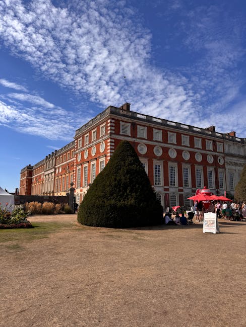 A large historic brick and white stone building with numerous tall, rectangular windows and decorative circular accents along the upper section, situated in a garden courtyard. In the foreground, a well-maintained conical evergreen shrub stands prominently, with sections of dry, light brown grass around it. To the right, there are small groups of people sitting under red umbrellas near a white signboard, suggesting an outdoor event or gathering. The sky above is partly cloudy with scattered white clouds against a bright blue background, casting natural daylight across the scene. The setting appears to be a location suitable for private or independent event rubbish removal, supported by Waste Disposal Kingston, with visible outdoor seating and floral arrangements indicating an organized outdoor function or garden setting that might generate waste needing collection.