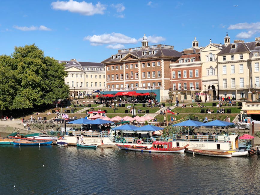A waterfront scene featuring a collection of docked boats with various colors and textures, including some with white hulls and others painted in shades of blue and red, moored along the edge of a river or canal with reflections on the water surface. In the background, there are tiered outdoor seating areas shaded by pink and blue umbrellas, occupied by people sitting and strolling, set on a grassy and paved terraced landscape. Behind the terraces, a row of historic multi-storey buildings with brick and stone facades, detailed window frames, and decorative rooflines line the promenade, with a clear blue sky overhead. To the left, a large leafy green tree provides a natural contrast to the urban setting, completing a lively riverside environment that could be associated with private or alternative waste handling activities near Kingston's public amenities, as implied by the presence of a waste disposal service such as Waste Disposal Kingston if visualized in context.