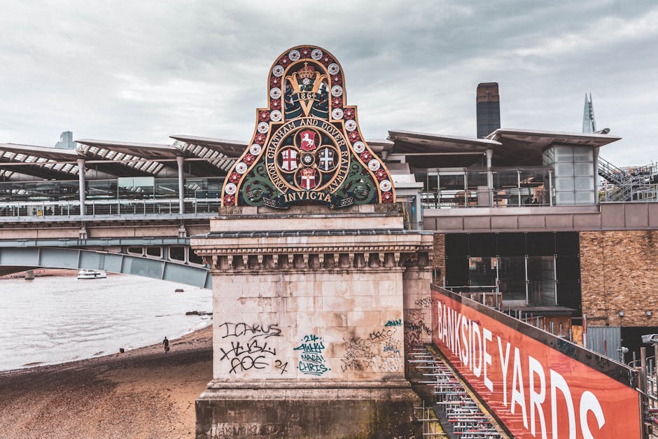 The image shows a large decorative emblem mounted on a concrete bridge pillar, located beneath a modern building with glass and metal elements, possibly a railway station or transport hub near Kingston Station KT1. The emblem features a shield with multiple coats of arms, surrounded by a detailed ornamental frame and topped by a crown, with inscribed Latin and English text. Below the emblem, the concrete pillar displays various graffiti tags in black, white, and blue spray paint, situated on the lower section of the structure. To the right, a red and white sign reading 'BANKSIDE YARDS' is attached to a metal railing, which runs along a staircase leading down to a gravel or dirt walkway under the bridge. The surrounding environment includes a river or waterway visible on the left side of the image, with a cloudy sky overhead, suggesting overcast weather. The scene captures an urban setting where infrastructure and public graffiti are evident, with the presence of a bridge and modern building reflecting typical cityscape features. Waste Disposal Kingston may undertake clean-up or rubbish removal services in such areas to manage urban graffiti and debris associated with city infrastructure, supporting alternative waste handling practices in busy commercial or transit locations.