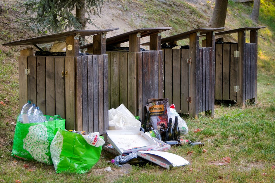 Four wooden outdoor trash bins with slanted roofs are positioned side by side on a grassy area, each constructed from vertical wooden planks with a dark weathered finish. The bins are situated on a patch of grass with scattered fallen leaves, and behind them, there is a natural environment featuring a gentle slope with grass, dirt, and a few trees. In front of the bins, discarded rubbish including two large green plastic shopping bags filled with plastic bottles and other waste, a white pizza box, and several glass bottles are scattered across the ground. Some of the waste is piled directly on the grass, with a few items leaning against the bins. The scene is illuminated by natural daylight, highlighting the contrast between the tidy appearance of the bins and the cluttered rubbish in front, illustrating an example of unmanaged waste disposal outside designated collection points, relevant to private or alternative rubbish handling services managed by Waste Disposal Kingston.