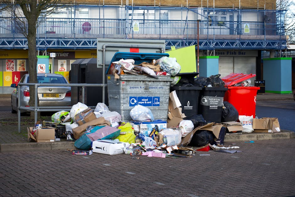 A roadside scene in front of a commercial area shows an overflowing waste disposal bin and numerous scattered rubbish bags, cardboard boxes, and loose waste items on a paved parking area. The large grey bin, designated for mixed paper and cardboard recycling, is visibly overfilled, with bulky paper waste and cardboard protruding from its open lid. Surrounding the bin are various black, red, and brown refuse sacks, some torn open, revealing contents such as plastic packaging, paper, and miscellaneous trash. Additional smaller cardboard boxes and loose items are strewn across the ground, contributing to a cluttered appearance. Behind this scene, there is a metal railing and parked cars, indicating a busy urban environment. In the background, a commercial building with a blue scaffolding system and signage is visible, with shopfronts beneath. The overall atmosphere suggests a scenario where private waste collection services, like those from Waste Disposal Kingston, have been used for an on-site rubbish removal, highlighting the importance of timely clearance to prevent accumulation and maintain cleanliness in busy commercial and public areas.